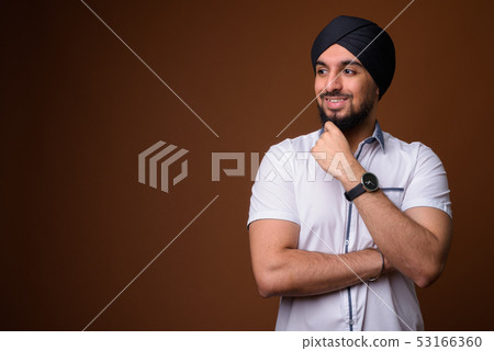 Young bearded Indian Sikh man wearing turban against brown backg 53166360