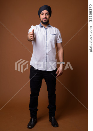 Young bearded Indian Sikh man wearing turban against brown backg 53166370