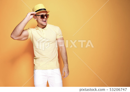 Confident stylish guy in yellow shirt, sunglasses, white shorts, straw hat looking side away from 53206747