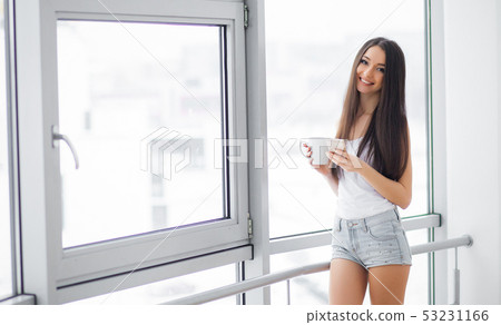 Happy morning. Portrait of beautiful young woman standing at window, drinking coffee and looking Happy morning. Portrait of beautiful young woman standing at window, drinking coffee and looking 53231166