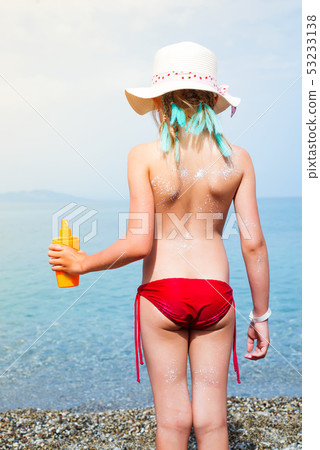 Little adorable girl with white hat and blue hair hold suncream bottle on the sea beach. Summer 53233138