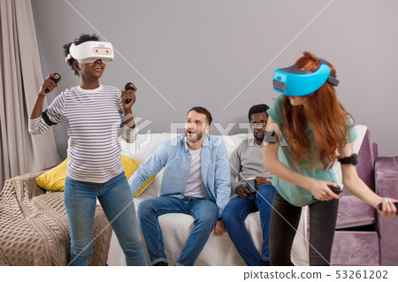Two multiracial young women dancing wearing virtual reality glasses while their boyfriends sitting 53261202