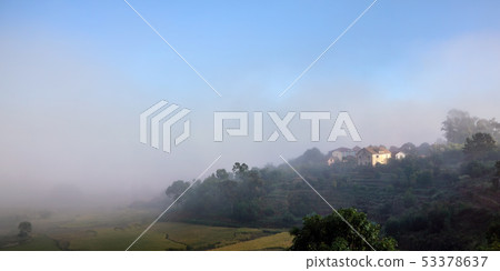 Morning fog over rice fields, sun shines through to small clay buildings - typical landscape in 53378637