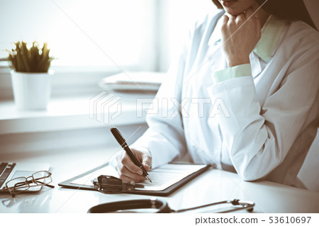 Doctor woman working with medication history records form while sitting at the desk near window in 53610697