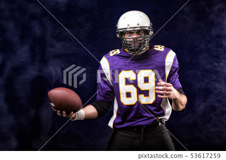 American football player holding ball in his hands in smoke. Black background, copy space. The American football player holding ball in his hands in smoke. Black background, copy space. The 53617259