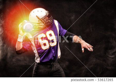 American football player holding ball in his hands in smoke. Black background, copy space. The American football player holding ball in his hands in smoke. Black background, copy space. The 53617265