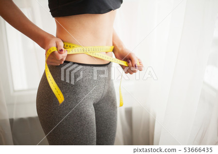 Hands measuring waist with a tape. Slim and healthy woman at her home Hands measuring waist with a tape. Slim and healthy woman at her home 53666435