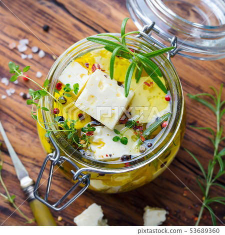 Feta cheese marinated in olive oil with fresh herbs in a glass jar. Wooden background. Top view. Feta cheese marinated in olive oil with fresh herbs in a glass jar. Wooden background. Top view. 53689360