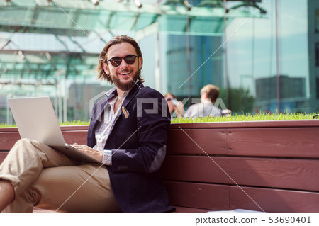 Photo of smiling male freelancer looking at camera with laptop in hands while sitting on wooden Photo of smiling male freelancer looking at camera with laptop in hands while sitting on wooden 53690401