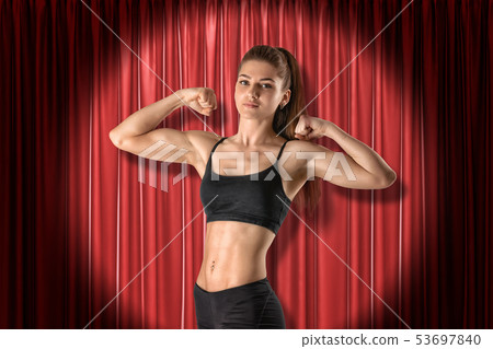 Crop image of young fit woman in crop top, standing in half-turn in spotlight at red stage curtain Crop image of young fit woman in crop top, standing in half-turn in spotlight at red stage curtain 53697840
