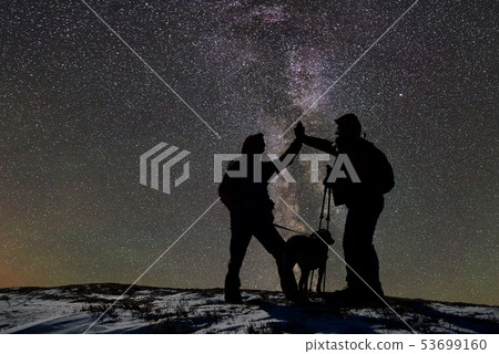 Silhouettes of two adult male skiers with dog on top of snow-covered mountain at night. Dark starry 53699160