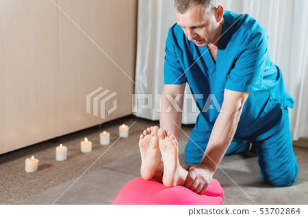 A male physiotherapist is stretching the knee joints to a young girl patient. Manual Wellness A male physiotherapist is stretching the knee joints to a young girl patient. Manual Wellness 53702864