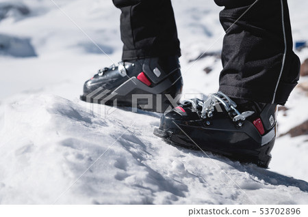 A close-up of a skier's leg in ski boots without skis stands on a knoll of bliss against the A close-up of a skier's leg in ski boots without skis stands on a knoll of bliss against the 53702896