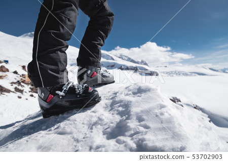A close-up of a skier's leg in ski boots without skis stands on a knoll of bliss against the A close-up of a skier's leg in ski boots without skis stands on a knoll of bliss against the 53702903