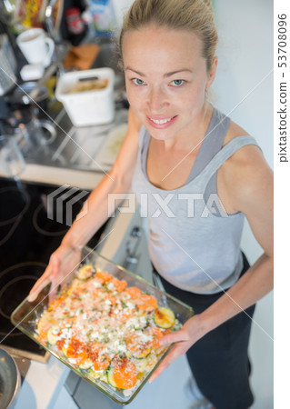 Smiling young healthy woman holding and proudly showing glass baking try with row vegetarian dish Smiling young healthy woman holding and proudly showing glass baking try with row vegetarian dish 53708096