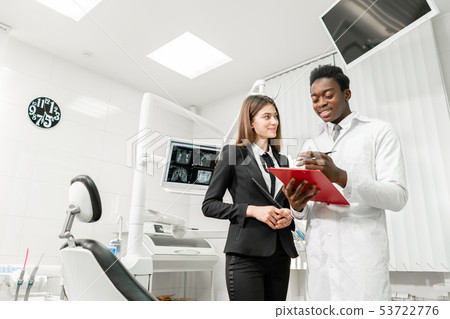 Woman patient in dental clinic. Young African male dentist makes a diagnosis and records Woman patient in dental clinic. Young African male dentist makes a diagnosis and records 53722776