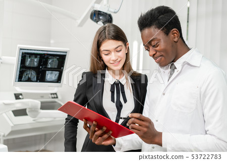 Young African male dentist makes a diagnosis and records recommendations. Woman patient at dental Young African male dentist makes a diagnosis and records recommendations. Woman patient at dental 53722783