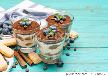 Classic tiramisu dessert in a glass with blueberries on blue wooden background Classic tiramisu dessert in a glass with blueberries on blue wooden background 53738517