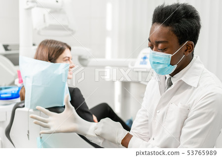 The doctor puts on rubber gloves. Young African male dentist with a patient. Woman in the dentist 53765989