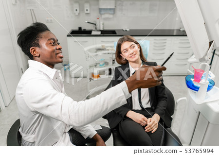 Young African male dentist shows x-rays on the monitor. Woman in the dentist chair at dental clinic Young African male dentist shows x-rays on the monitor. Woman in the dentist chair at dental clinic 53765994