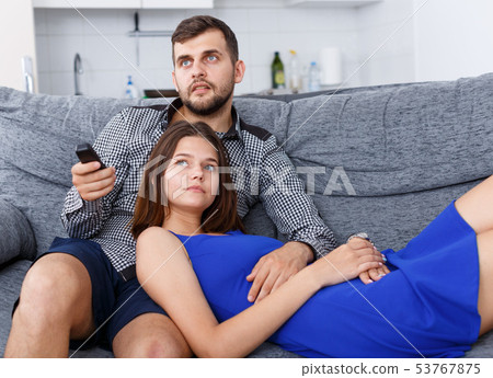 Cheerful young couple with TV remote sitting at sofa watching movie 53767875