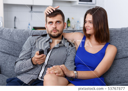 Cheerful young couple with TV remote sitting at sofa watching movie Cheerful young couple with TV remote sitting at sofa watching movie 53767877