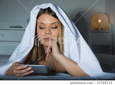 Young female under blanket on bed with smartphone at home Young female under blanket on bed with smartphone at home 53768714
