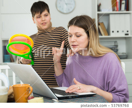 Mother working at laptop and unhappy son standing racquets for tennis Mother working at laptop and unhappy son standing racquets for tennis 53769768