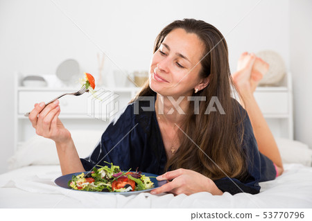 Young pretty woman eating vegetable salad from plato in bed Young pretty woman eating vegetable salad from plato in bed 53770796