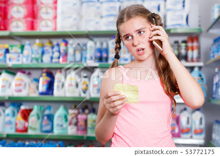 active girl standing in store choosing products while talking by phone and looking at shopping list 53772175