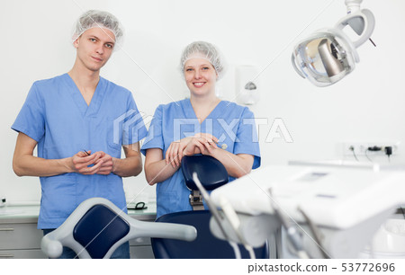 Two professional confident dentists posing near dental chair in clinic Two professional confident dentists posing near dental chair in clinic 53772696
