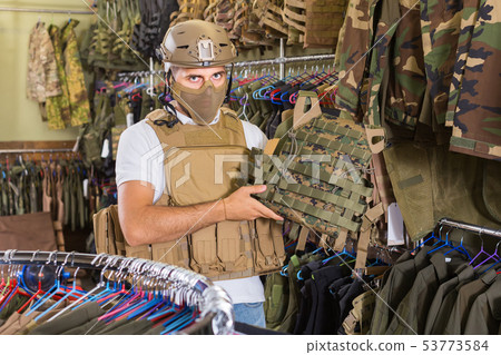 Young guy choosing flak jacket in military shop Young guy choosing flak jacket in military shop 53773584