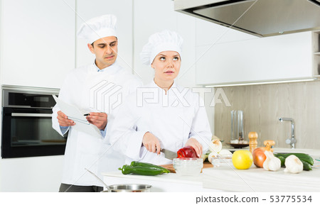 Cheerful woman and man young chefs preparing food with paper recipe Cheerful woman and man young chefs preparing food with paper recipe 53775534