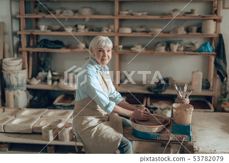 Elderly craftswoman making clay object while sitting in potter's studio 53782079