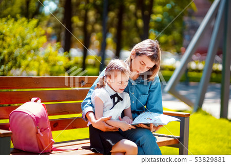 Mother and little daughter in outdoors public park sitting on the bench and read book, study lessons 53829881