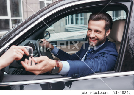 Happy and successful. Cheerful and stylish young businessman in full suit is taking a key from car Happy and successful. Cheerful and stylish young businessman in full suit is taking a key from car 53858242