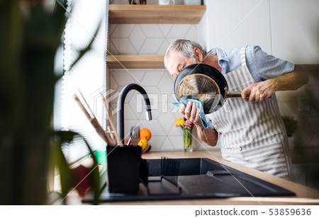 A senior man indoors in kitchen at home, washing up a pan. 53859636