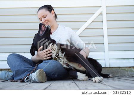 Photo of sitting woman in jeans and white jacket with black dog on background of white wooden wall Photo of sitting woman in jeans and white jacket with black dog on background of white wooden wall 53882510