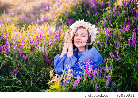 Young pretty woman with flower wreath sitting in purple field. Portrait of girl in blue clothing. Young pretty woman with flower wreath sitting in purple field. Portrait of girl in blue clothing. 53889931