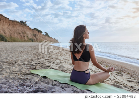 Happy young woman practices yoga and meditates in the lotus position on the beach Happy young woman practices yoga and meditates in the lotus position on the beach 53894488