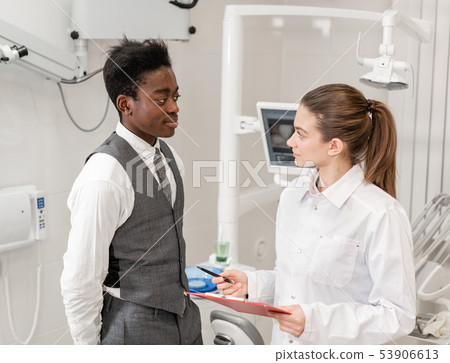 African male patient in dental clinic. Young Woman dentist makes a diagnosis and records African male patient in dental clinic. Young Woman dentist makes a diagnosis and records 53906613