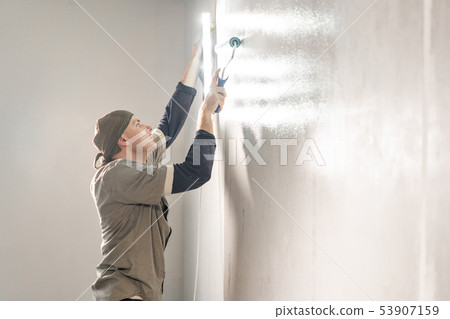 Young repairman smear on wall glue with a roller. . Worker glueing wallpapers on concrete wall 53907159