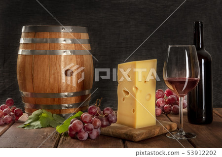 A glass of red wine with a bottle, a wine barrel, a large piece of cheesse, grapes, and vine leaves 53912202