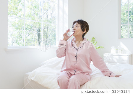 Senior woman in pajamas drinking water on bed Senior woman in pajamas drinking water on bed 53945124