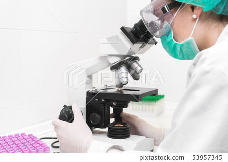 Young female doctor working with a microscope in a laboratory, protected against biohazard with mask 53957345