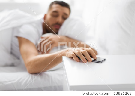 sleepy young man reaching for smartphone in bed sleepy young man reaching for smartphone in bed 53985265