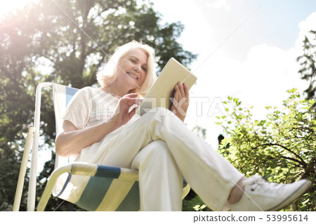 Senior blonde woman sitting in chair and using digital tablet in garden Senior blonde woman sitting in chair and using digital tablet in garden 53995614