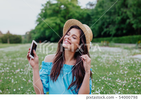 Beautiful woman in straw hat listening to music with headphones and sitting in a garden of white 54000798