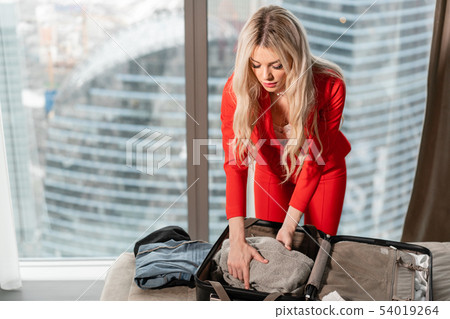 Young blond businesswoman arrives in a hotel room with black suitcase. Woman in red coral business Young blond businesswoman arrives in a hotel room with black suitcase. Woman in red coral business 54019264