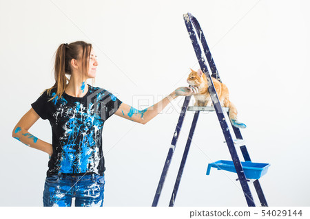 Young woman painter stroking a cat, designer and worker paints the wall. The pet sits next on the Young woman painter stroking a cat, designer and worker paints the wall. The pet sits next on the 54029144